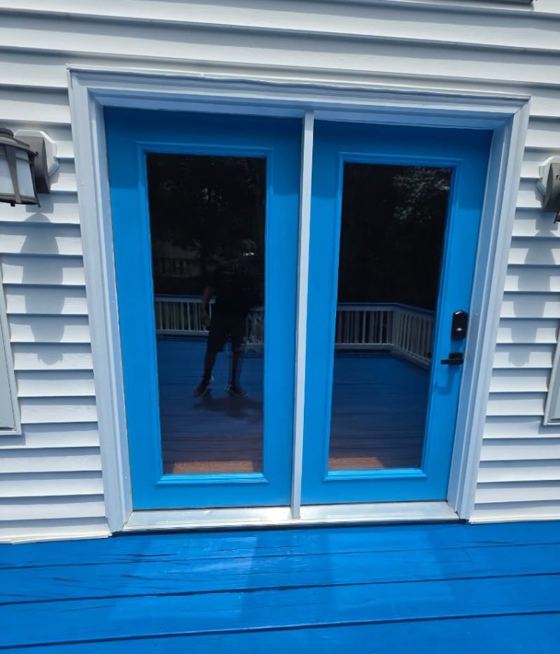 Deck staining project featuring a bright blue wooden deck with matching blue patio doors, glass panels, white trim, and exterior siding.