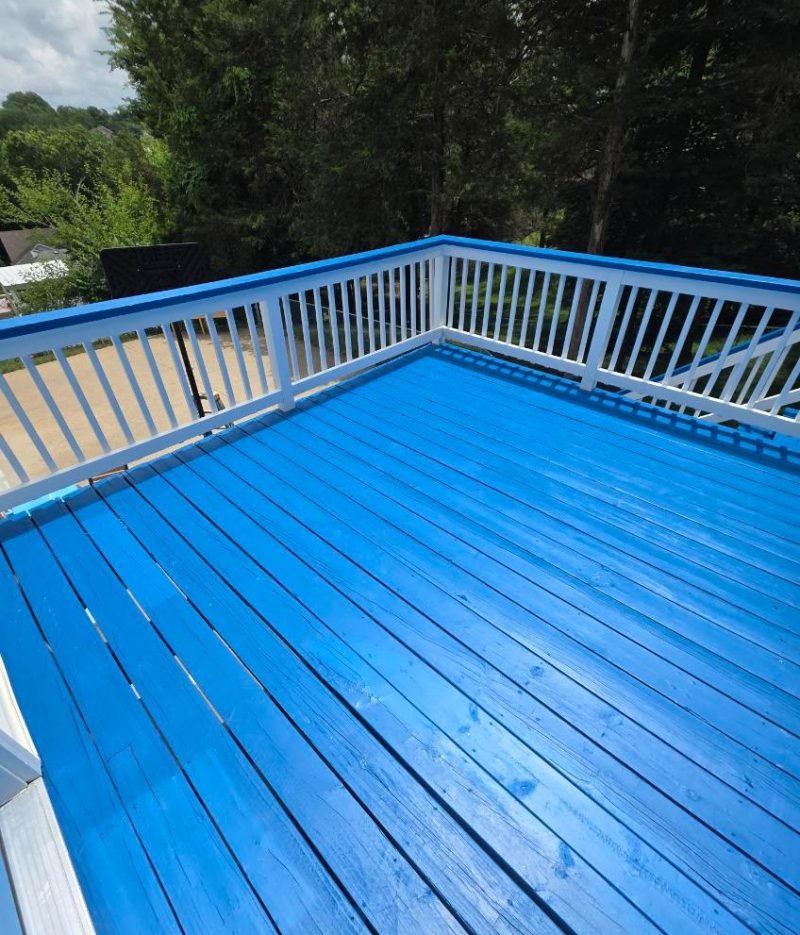 Deck staining project featuring a bright blue wooden deck with white railings, outdoor seating, and trees in the background.