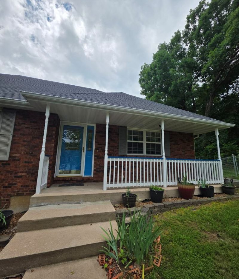 Door staining project featuring a blue front door on a brick home with a covered porch, white railing, front steps, and potted plants.