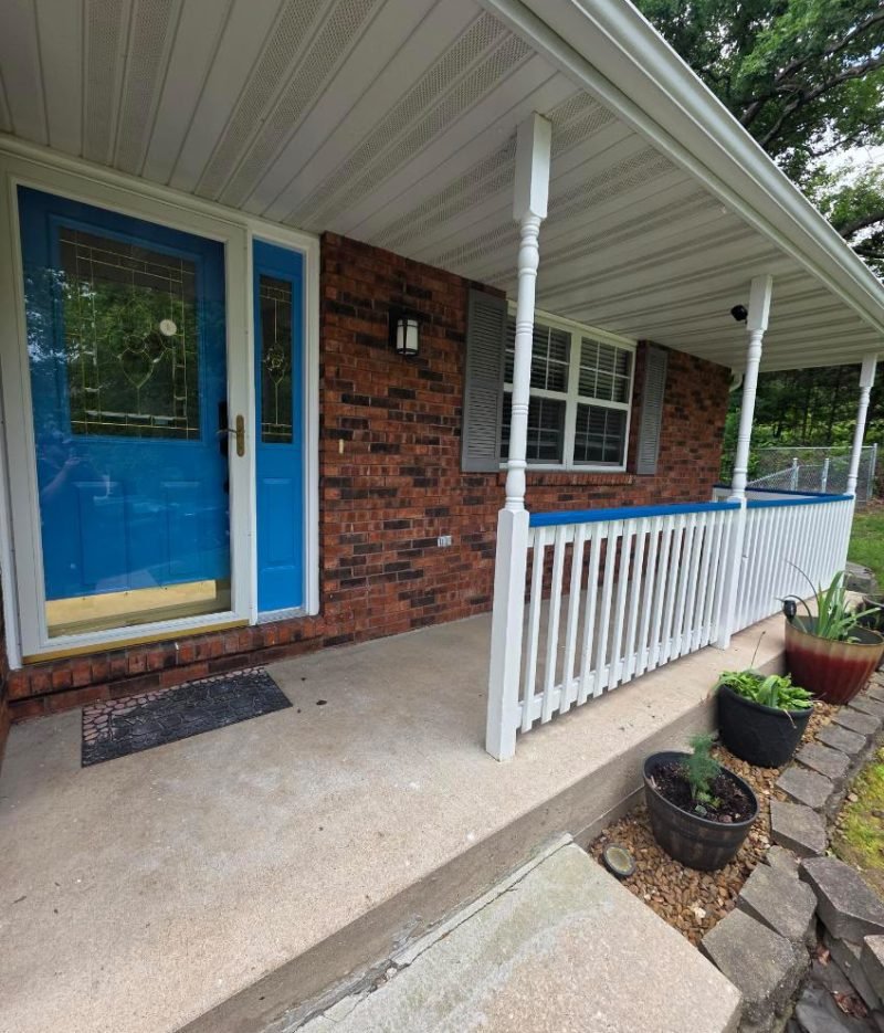 Door staining service on a bright blue front door with decorative glass panels, white porch railing, brick exterior, and potted plants.
